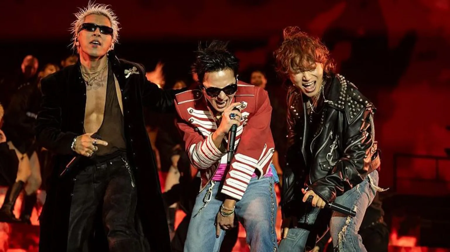 Timothy Norris/Getty Images Three performers stand at the front of a stage holding handheld microphones during a live performance. One performer wears a long black coat and sunglasses, another wears a red military-style jacket with white trim and jeans, and a third wears a black studded leather jacket and ripped jeans. The stage is lit in red, with flames or flame-effect visuals displayed behind the performers.